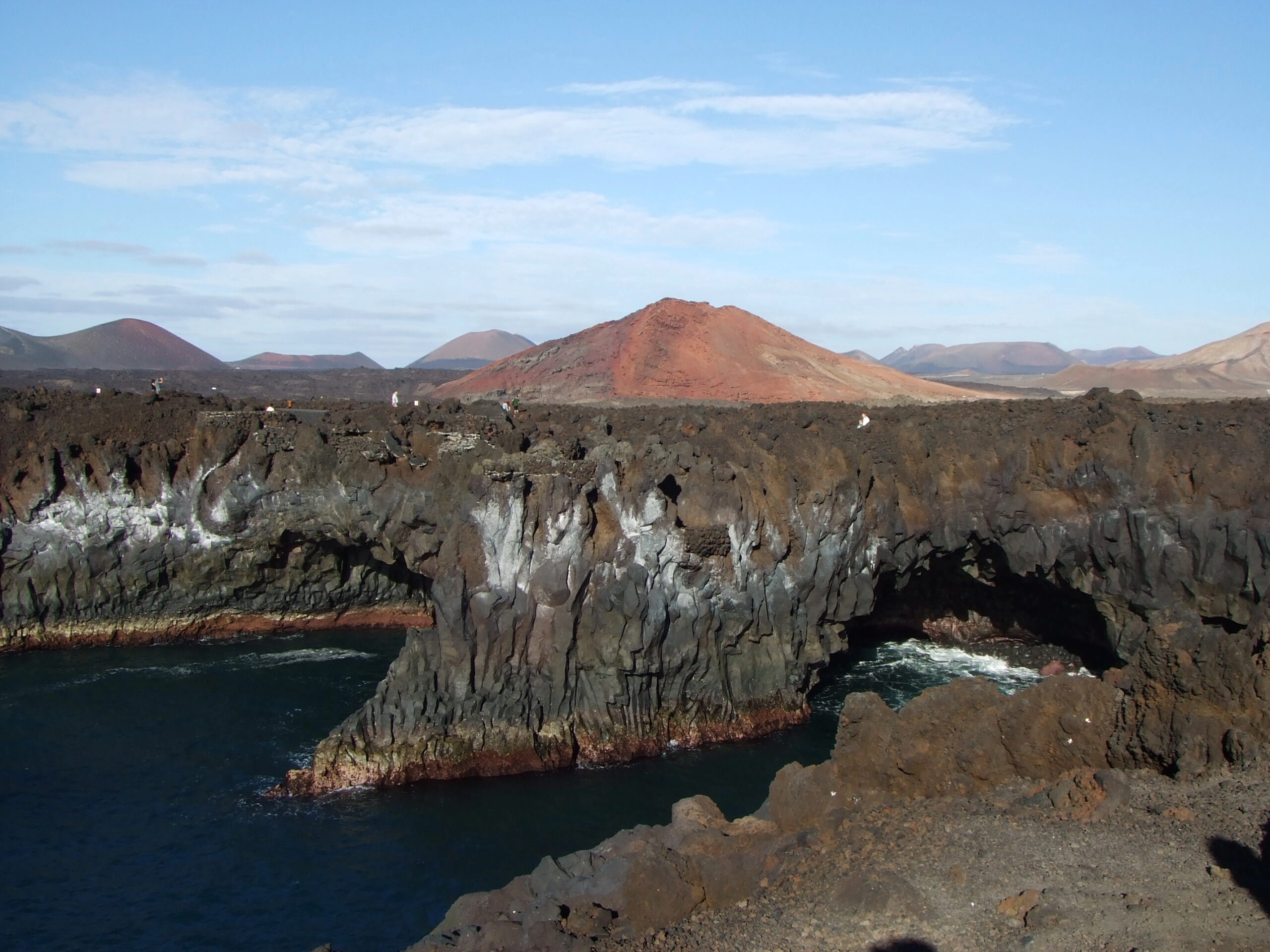 Vulkanische kliffen en natuurlijke rotsbogen van Los Hervideros aan de westkust van Lanzarote met uitzicht op de Atlantische Oceaan en de rode vulkaanbergen van het Timanfaya-gebied