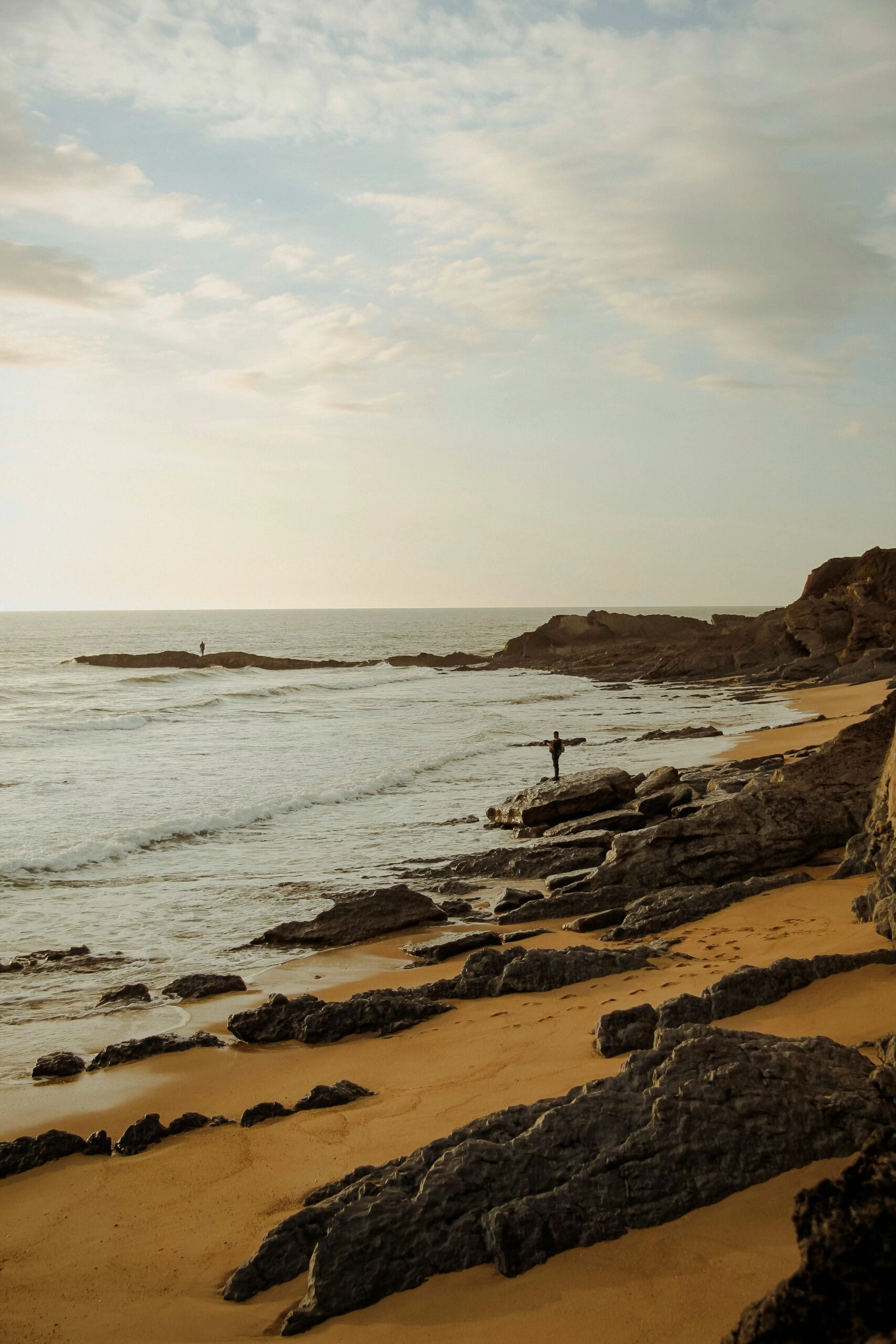 Gouden zandstrand met rotsen en golven aan de Atlantische kust bij Praia de Mira in Portugal, met een wandelaar aan zee tijdens zonsondergang.