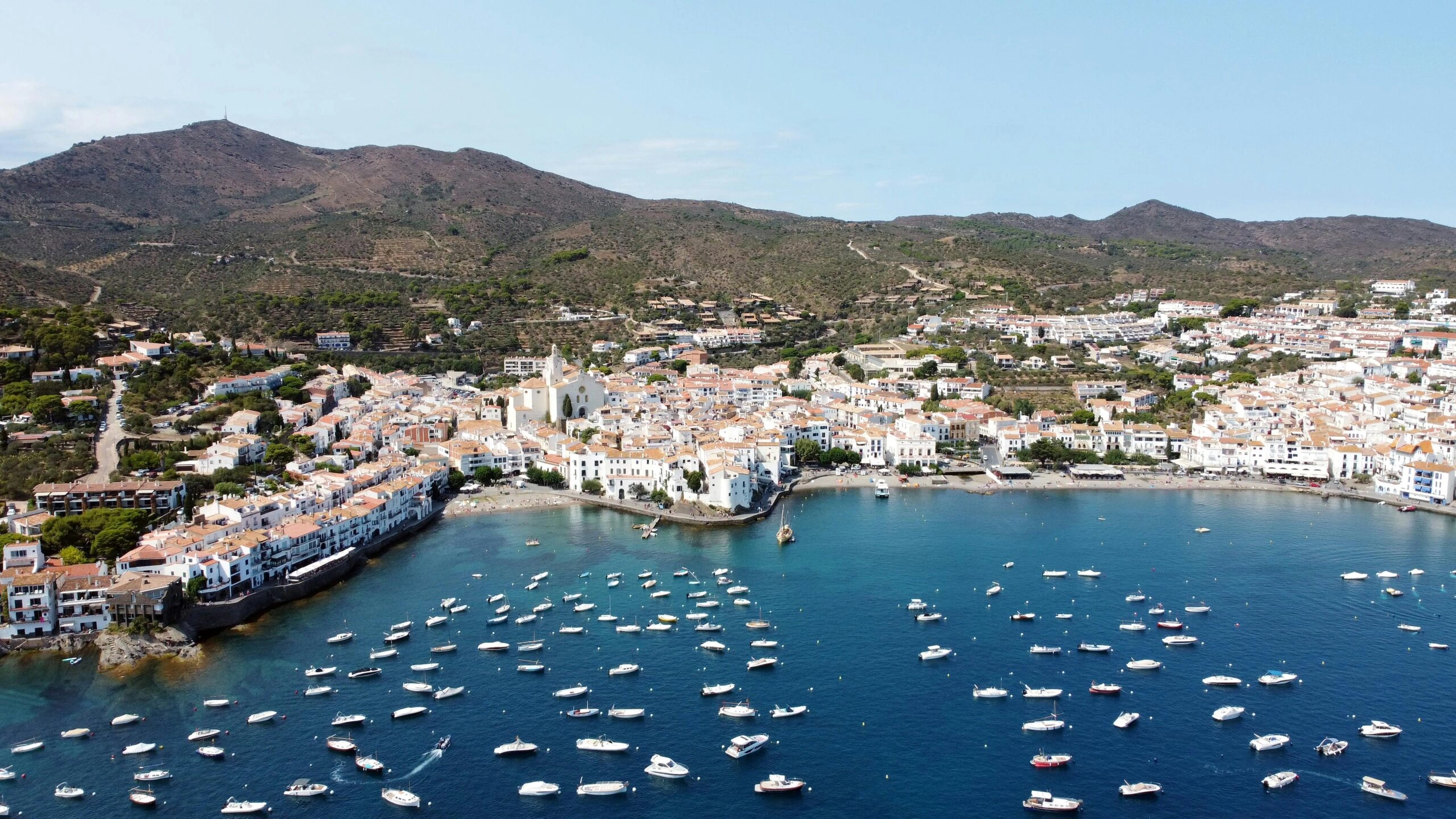 Luchtfoto van het witte kustdorp Cadaqués met baai vol bootjes en heuvels op de achtergrond.