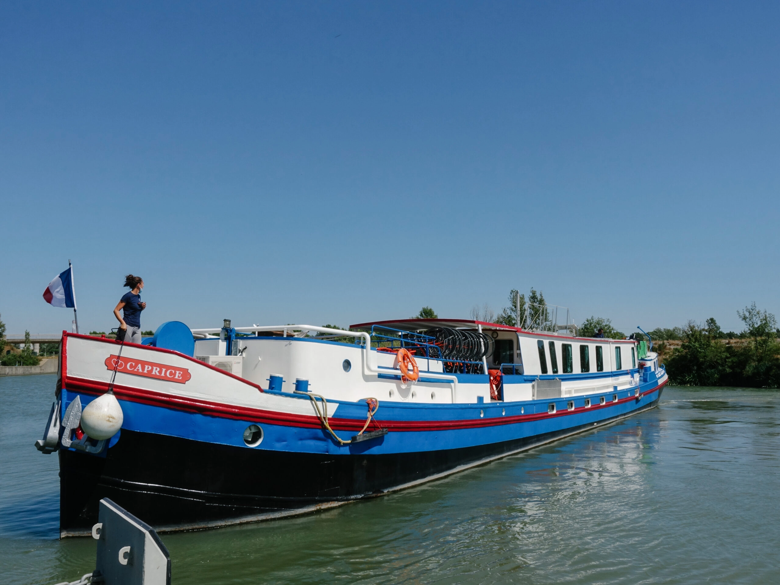 De riviercruiseboot Caprice vaart door een kanaal in de Camargue, met fietsen aan dek en een bemanningslid aan de voorzijde.