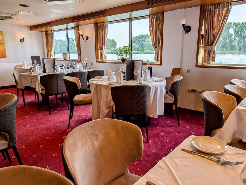 Restaurant van MS Olympia met gedekte tafels en grote panoramavensters met uitzicht over de rivier.