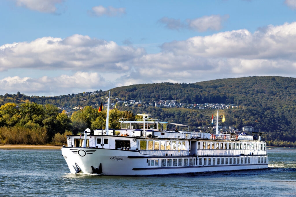 MS Olympia riviercruiseschip varend over de Rijn met groene heuvels en dorpen op de achtergrond, ideaal voor fiets- en vaarvakanties.
