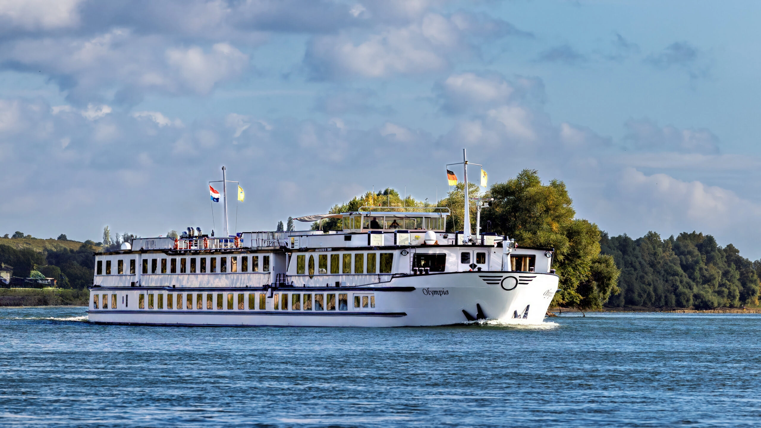Riviercruiseschip MS Olympia varend over de Rijn met zonnedek, panoramaramen en groene rivieroevers op de achtergrond.